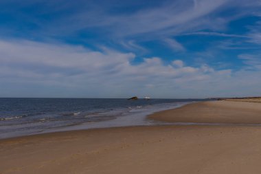 Uzak beyaz tekne ve kara gemi kalıntıları parçalı bulutlu bir gökyüzünün altında engin, koyu mavi bir denizde oturuyorlar. Ön planda Sandy Shore var. Anımsatıcı kıyı manzarası.