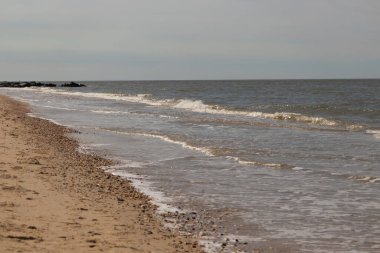 Yumuşak dalgalar, berrak ve solgun bir gökyüzünün altında kumlu ve çakıllı bir plaja yuvarlanıyor. Huzurlu sahil manzarası. Kum ve suyun doğal dokuları.