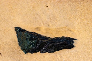 Koyu, parıldayan bir deniz kömürü ya da antrasit parçası açık kumların üzerinde yatar. Karşıtlık, hem kömürün hem de plajın dokusunu vurguluyor..