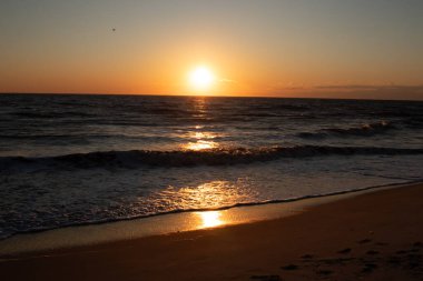 Güneşin ufukta battığı nefes kesici bir okyanus gün batımı dalgalanan mavi suya sıcak bir altın parıltı saçıyor. Huzurlu ve resimli sahil manzarası.