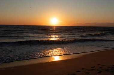 Güneşin ufukta battığı nefes kesici bir okyanus gün batımı dalgalanan mavi suya sıcak bir altın parıltı saçıyor. Huzurlu ve resimli sahil manzarası.