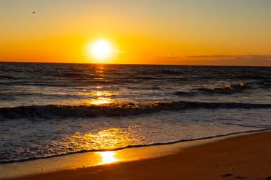 Güneşin ufukta battığı nefes kesici bir okyanus gün batımı dalgalanan mavi suya sıcak bir altın parıltı saçıyor. Huzurlu ve resimli sahil manzarası.