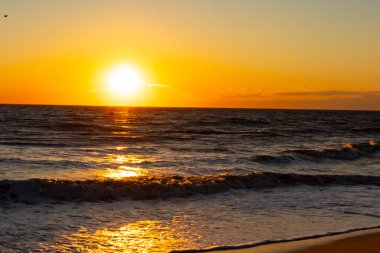 Güneşin ufukta battığı nefes kesici bir okyanus gün batımı dalgalanan mavi suya sıcak bir altın parıltı saçıyor. Huzurlu ve resimli sahil manzarası.