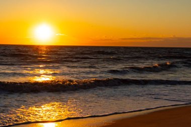 Güneşin ufukta battığı nefes kesici bir okyanus gün batımı dalgalanan mavi suya sıcak bir altın parıltı saçıyor. Huzurlu ve resimli sahil manzarası.