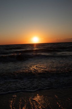 Güneşin ufukta battığı nefes kesici bir okyanus gün batımı dalgalanan mavi suya sıcak bir altın parıltı saçıyor. Huzurlu ve resimli sahil manzarası.