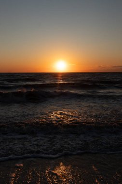 Güneşin ufukta battığı nefes kesici bir okyanus gün batımı dalgalanan mavi suya sıcak bir altın parıltı saçıyor. Huzurlu ve resimli sahil manzarası.