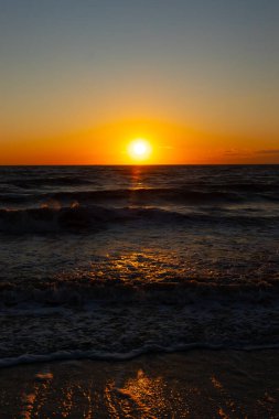 Güneşin ufukta battığı nefes kesici bir okyanus gün batımı dalgalanan mavi suya sıcak bir altın parıltı saçıyor. Huzurlu ve resimli sahil manzarası.