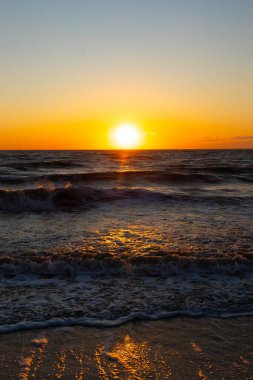 Güneşin ufukta battığı nefes kesici bir okyanus gün batımı dalgalanan mavi suya sıcak bir altın parıltı saçıyor. Huzurlu ve resimli sahil manzarası.