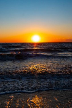 Güneşin ufukta battığı nefes kesici bir okyanus gün batımı dalgalanan mavi suya sıcak bir altın parıltı saçıyor. Huzurlu ve resimli sahil manzarası.