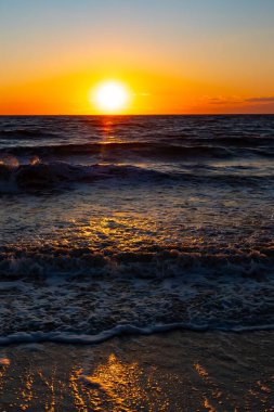 Güneşin ufukta battığı nefes kesici bir okyanus gün batımı dalgalanan mavi suya sıcak bir altın parıltı saçıyor. Huzurlu ve resimli sahil manzarası.