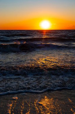 Güneşin ufukta battığı nefes kesici bir okyanus gün batımı dalgalanan mavi suya sıcak bir altın parıltı saçıyor. Huzurlu ve resimli sahil manzarası.