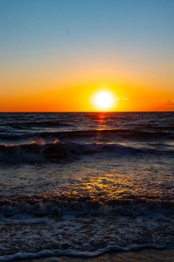 Güneşin ufukta battığı nefes kesici bir okyanus gün batımı dalgalanan mavi suya sıcak bir altın parıltı saçıyor. Huzurlu ve resimli sahil manzarası.