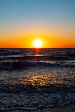 Güneşin ufukta battığı nefes kesici bir okyanus gün batımı dalgalanan mavi suya sıcak bir altın parıltı saçıyor. Huzurlu ve resimli sahil manzarası.