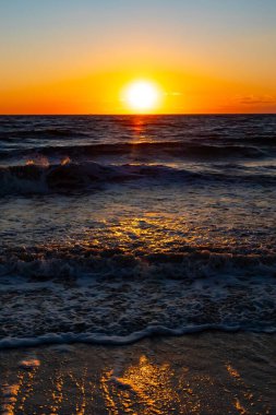 Güneşin ufukta battığı nefes kesici bir okyanus gün batımı dalgalanan mavi suya sıcak bir altın parıltı saçıyor. Huzurlu ve resimli sahil manzarası.
