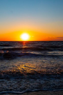 Güneşin ufukta battığı nefes kesici bir okyanus gün batımı dalgalanan mavi suya sıcak bir altın parıltı saçıyor. Huzurlu ve resimli sahil manzarası.