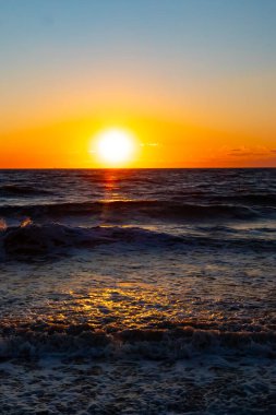 Güneşin ufukta battığı nefes kesici bir okyanus gün batımı dalgalanan mavi suya sıcak bir altın parıltı saçıyor. Huzurlu ve resimli sahil manzarası.