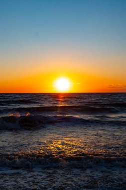 Güneşin ufukta battığı nefes kesici bir okyanus gün batımı dalgalanan mavi suya sıcak bir altın parıltı saçıyor. Huzurlu ve resimli sahil manzarası.