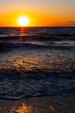 Güneşin ufukta battığı nefes kesici bir okyanus gün batımı dalgalanan mavi suya sıcak bir altın parıltı saçıyor. Huzurlu ve resimli sahil manzarası.