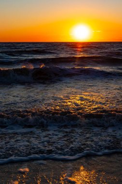 Güneşin ufukta battığı nefes kesici bir okyanus gün batımı dalgalanan mavi suya sıcak bir altın parıltı saçıyor. Huzurlu ve resimli sahil manzarası.