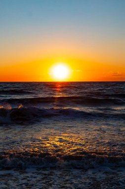 Güneşin ufukta battığı nefes kesici bir okyanus gün batımı dalgalanan mavi suya sıcak bir altın parıltı saçıyor. Huzurlu ve resimli sahil manzarası.