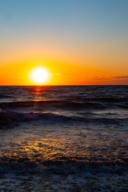 Güneşin ufukta battığı nefes kesici bir okyanus gün batımı dalgalanan mavi suya sıcak bir altın parıltı saçıyor. Huzurlu ve resimli sahil manzarası.