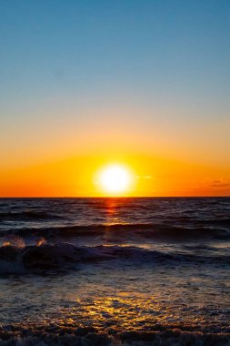 Güneşin ufukta battığı nefes kesici bir okyanus gün batımı dalgalanan mavi suya sıcak bir altın parıltı saçıyor. Huzurlu ve resimli sahil manzarası.