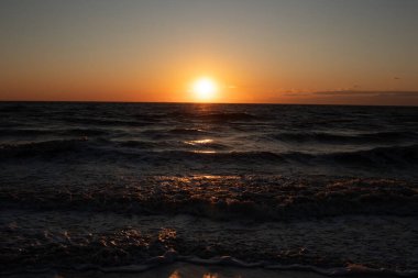 Güneşin ufukta battığı nefes kesici bir okyanus gün batımı dalgalanan mavi suya sıcak bir altın parıltı saçıyor. Huzurlu ve resimli sahil manzarası.