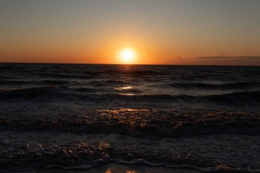 Güneşin ufukta battığı nefes kesici bir okyanus gün batımı dalgalanan mavi suya sıcak bir altın parıltı saçıyor. Huzurlu ve resimli sahil manzarası.