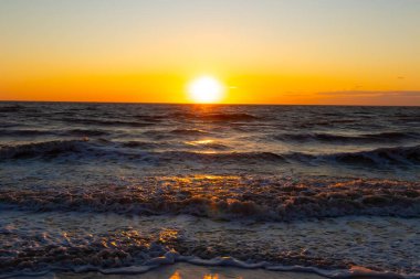 Güneşin ufukta battığı nefes kesici bir okyanus gün batımı dalgalanan mavi suya sıcak bir altın parıltı saçıyor. Huzurlu ve resimli sahil manzarası.