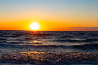 Güneşin ufukta battığı nefes kesici bir okyanus gün batımı dalgalanan mavi suya sıcak bir altın parıltı saçıyor. Huzurlu ve resimli sahil manzarası.