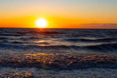Güneşin ufukta battığı nefes kesici bir okyanus gün batımı dalgalanan mavi suya sıcak bir altın parıltı saçıyor. Huzurlu ve resimli sahil manzarası.