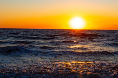 Güneşin ufukta battığı nefes kesici bir okyanus gün batımı dalgalanan mavi suya sıcak bir altın parıltı saçıyor. Huzurlu ve resimli sahil manzarası.