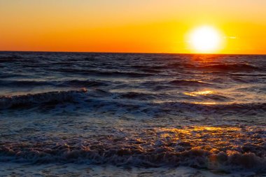Güneşin ufukta battığı nefes kesici bir okyanus gün batımı dalgalanan mavi suya sıcak bir altın parıltı saçıyor. Huzurlu ve resimli sahil manzarası.