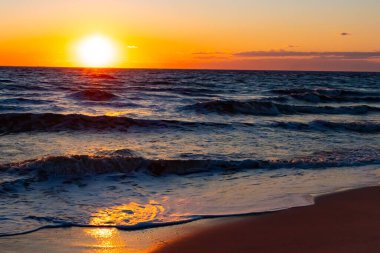 Güneşin ufukta battığı nefes kesici bir okyanus gün batımı dalgalanan mavi suya sıcak bir altın parıltı saçıyor. Huzurlu ve resimli sahil manzarası.