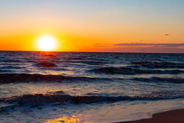 Güneşin ufukta battığı nefes kesici bir okyanus gün batımı dalgalanan mavi suya sıcak bir altın parıltı saçıyor. Huzurlu ve resimli sahil manzarası.