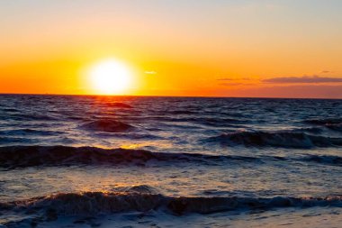 Güneşin ufukta battığı nefes kesici bir okyanus gün batımı dalgalanan mavi suya sıcak bir altın parıltı saçıyor. Huzurlu ve resimli sahil manzarası.