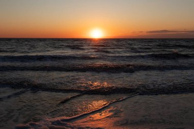 Güneşin ufukta battığı nefes kesici bir okyanus gün batımı dalgalanan mavi suya sıcak bir altın parıltı saçıyor. Huzurlu ve resimli sahil manzarası.