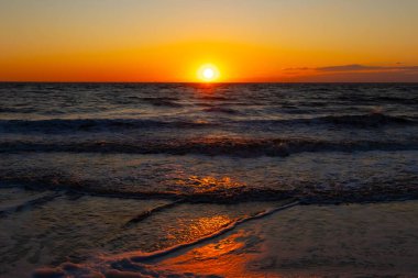 Güneşin ufukta battığı nefes kesici bir okyanus gün batımı dalgalanan mavi suya sıcak bir altın parıltı saçıyor. Huzurlu ve resimli sahil manzarası.