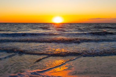 Güneşin ufukta battığı nefes kesici bir okyanus gün batımı dalgalanan mavi suya sıcak bir altın parıltı saçıyor. Huzurlu ve resimli sahil manzarası.