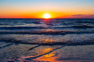 Güneşin ufukta battığı nefes kesici bir okyanus gün batımı dalgalanan mavi suya sıcak bir altın parıltı saçıyor. Huzurlu ve resimli sahil manzarası.