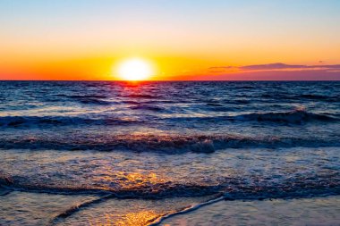 Güneşin ufukta battığı nefes kesici bir okyanus gün batımı dalgalanan mavi suya sıcak bir altın parıltı saçıyor. Huzurlu ve resimli sahil manzarası.