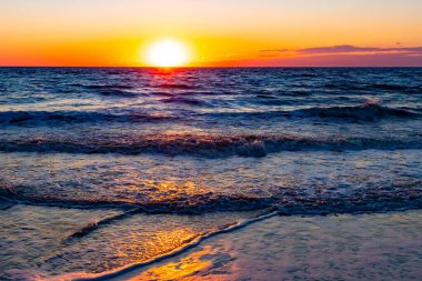 Güneşin ufukta battığı nefes kesici bir okyanus gün batımı dalgalanan mavi suya sıcak bir altın parıltı saçıyor. Huzurlu ve resimli sahil manzarası.