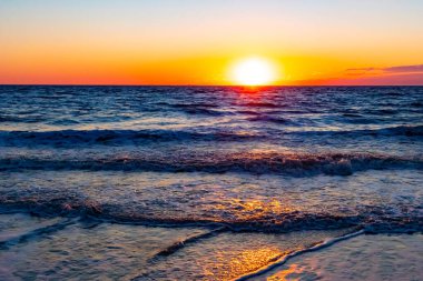 Güneşin ufukta battığı nefes kesici bir okyanus gün batımı dalgalanan mavi suya sıcak bir altın parıltı saçıyor. Huzurlu ve resimli sahil manzarası.