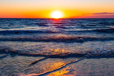 Güneşin ufukta battığı nefes kesici bir okyanus gün batımı dalgalanan mavi suya sıcak bir altın parıltı saçıyor. Huzurlu ve resimli sahil manzarası.