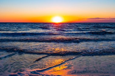 Güneşin ufukta battığı nefes kesici bir okyanus gün batımı dalgalanan mavi suya sıcak bir altın parıltı saçıyor. Huzurlu ve resimli sahil manzarası.