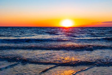 Güneşin ufukta battığı nefes kesici bir okyanus gün batımı dalgalanan mavi suya sıcak bir altın parıltı saçıyor. Huzurlu ve resimli sahil manzarası.