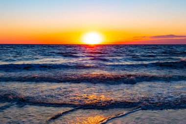 Güneşin ufukta battığı nefes kesici bir okyanus gün batımı dalgalanan mavi suya sıcak bir altın parıltı saçıyor. Huzurlu ve resimli sahil manzarası.