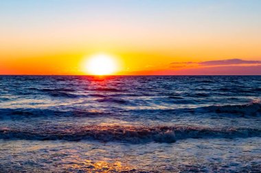 Güneşin ufukta battığı nefes kesici bir okyanus gün batımı dalgalanan mavi suya sıcak bir altın parıltı saçıyor. Huzurlu ve resimli sahil manzarası.
