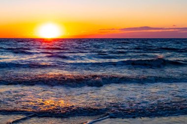 Güneşin ufukta battığı nefes kesici bir okyanus gün batımı dalgalanan mavi suya sıcak bir altın parıltı saçıyor. Huzurlu ve resimli sahil manzarası.