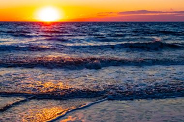 Güneşin ufukta battığı nefes kesici bir okyanus gün batımı dalgalanan mavi suya sıcak bir altın parıltı saçıyor. Huzurlu ve resimli sahil manzarası.